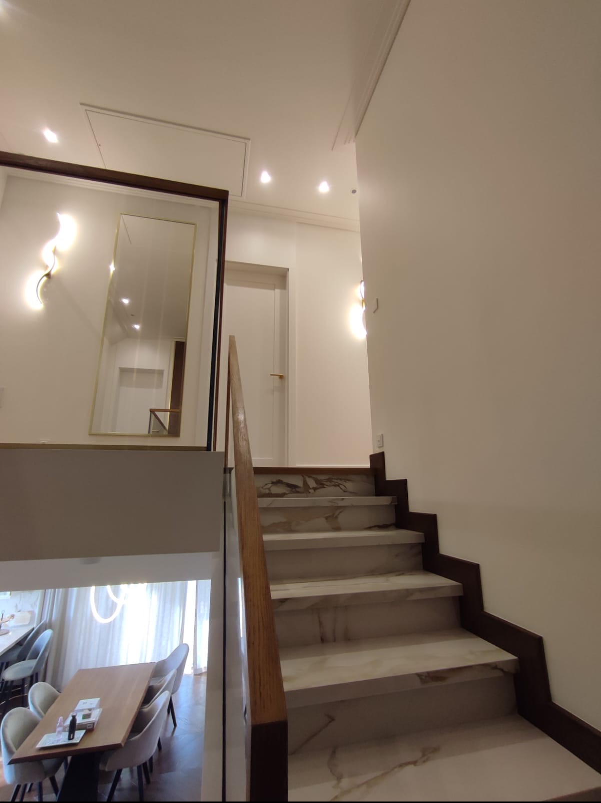 Schody wyłożone marmurem z drewnianą balustradą i szklaną barierką, widok na jadalnię z białymi krzesłami i drewnianym stołem, duże lustro w ciemnej ramie na ścianie.