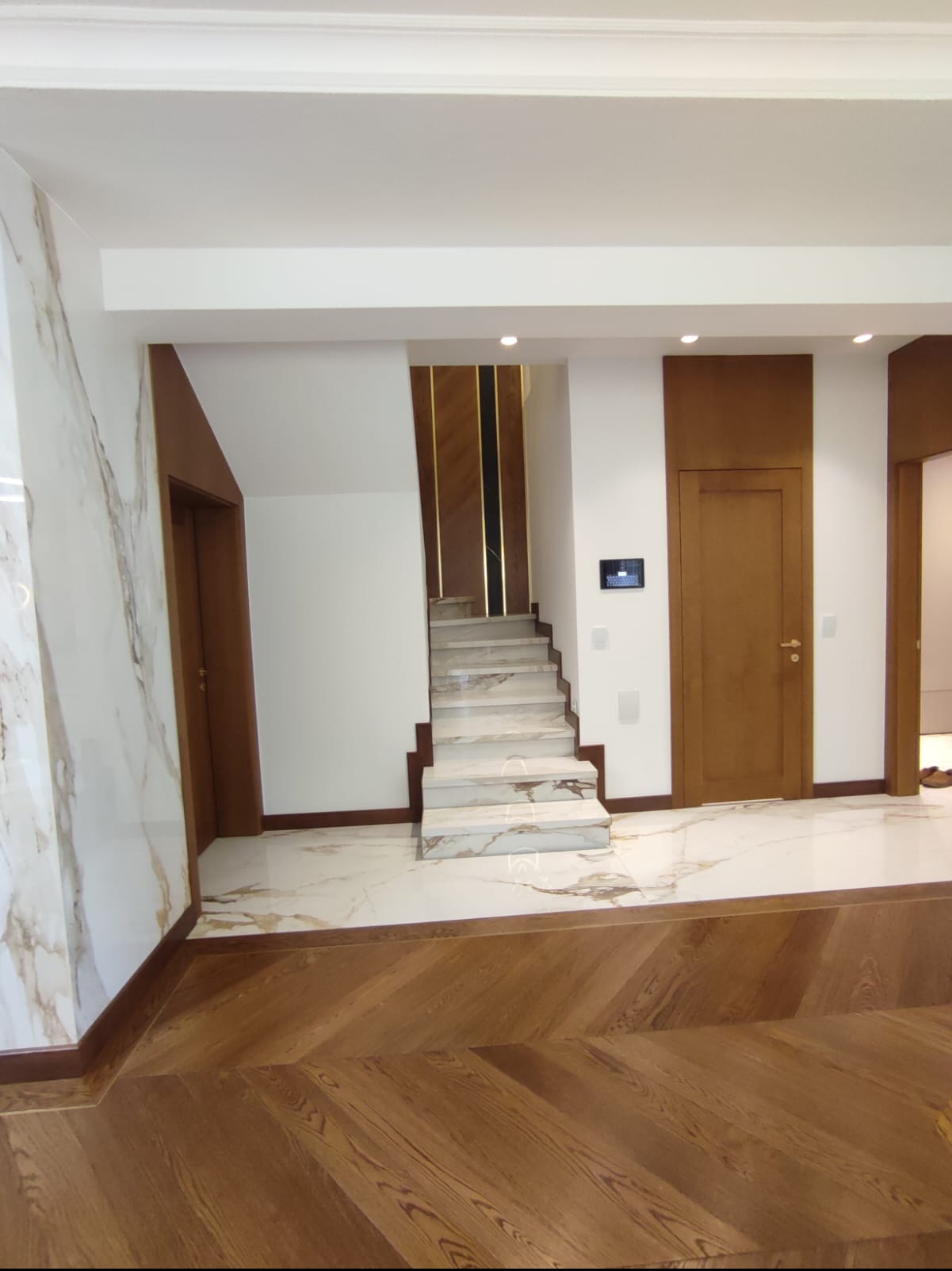 Korytarz z marmurowymi płytkami podłogowymi i schodami, drewnianą podłogą w jodełkę i dwiema parami drzwi z drewnianymi framugami.