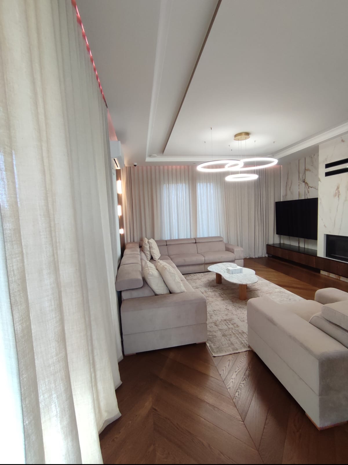 Jasne, przestronne wnętrze salonu z parkietem w jodełkę, beżową sofą narożną, marmurowym kominkiem i nowoczesnym oświetleniem.