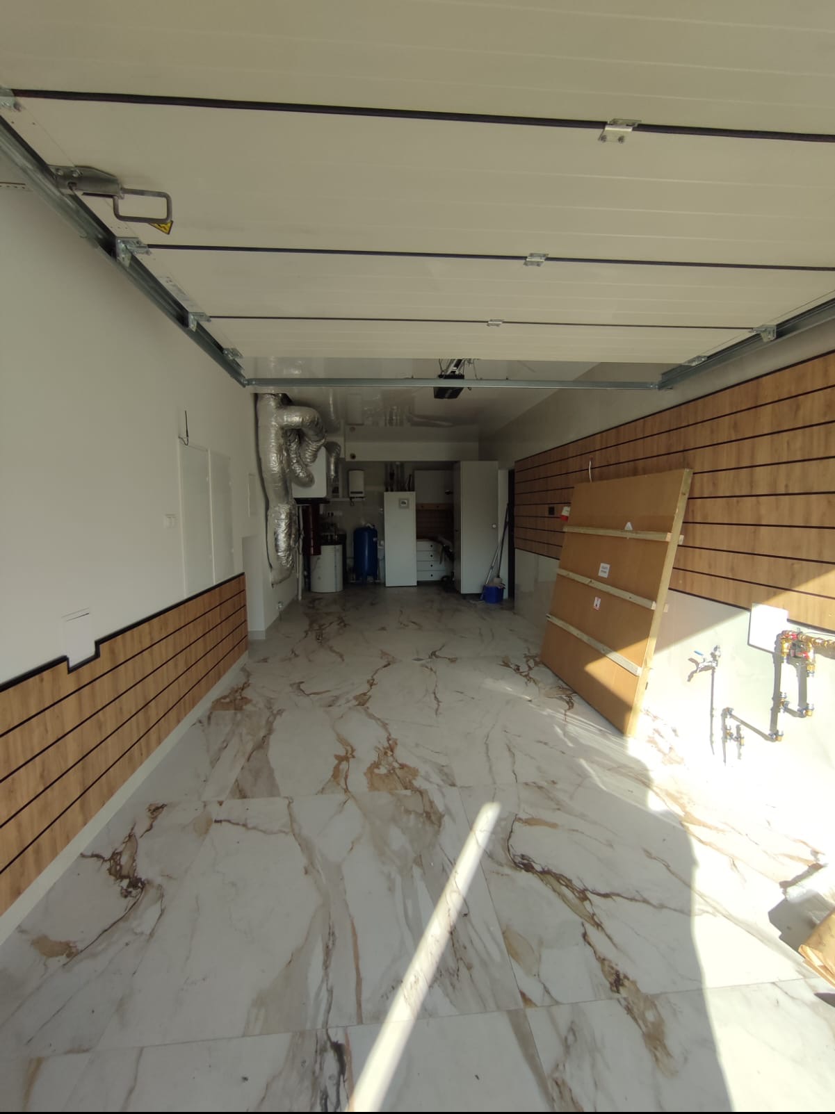 Wnętrze garażu po remoncie z białymi ścianami, panelami drewnopodobnymi na ścianach do połowy wysokości, marmurową posadzką i instalacją grzewczą w tle.