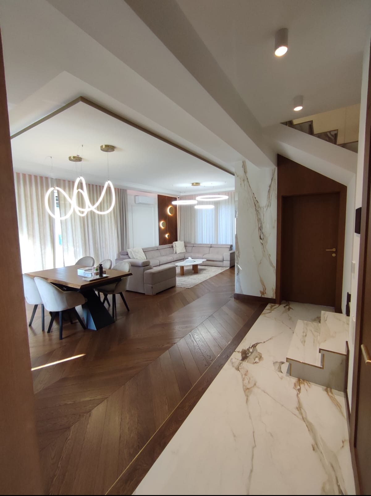 Przestronne wnętrze domu z parkietem w jodełkę, marmurowymi płytkami na schodach i ścianie, nowoczesnym oświetleniem i beżową sofą narożną.