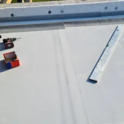 Montaż hydroizolacji na płaskim dachu: widoczna membrana, poziomica, młotek, wkrętarka i pudełko z mocowaniami.