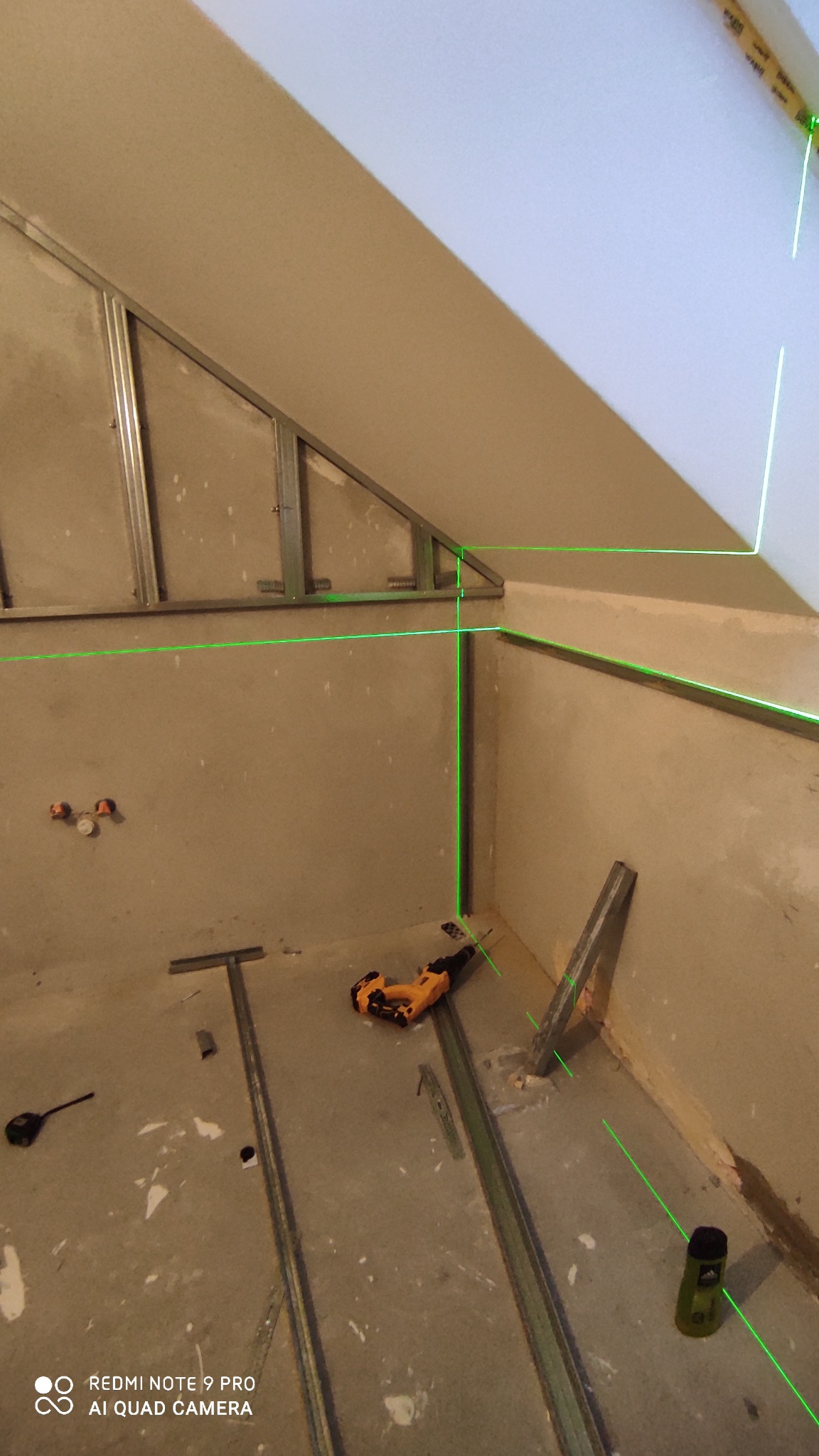 Szkielet konstrukcji metalowej pod płytę gipsowo-kartonową na skośnej ścianie poddasza, wyznaczony laserowo, z widocznymi narzędziami i elementami montażowymi na podłodze.