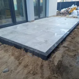 Wyłożony płytami taras z obrzeżem z ciemnej kostki, otoczony piaskiem, widok na przesuwne drzwi tarasowe i fragment zagospodarowanego terenu z tyłu.