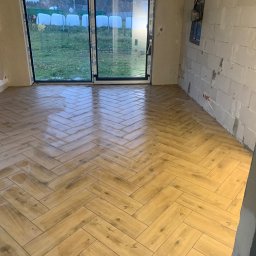 USŁUGI OGÓLNOBUDOWLANE RT - Ułożona pod kątem jasnobrązowa podłoga z płytek imitujących drewno w surowym pomieszczeniu z niezakończonymi ścianami i dużym oknem balkonowym.