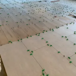 Układanie podłogi z płytek w dwóch kolorach, z użyciem systemu poziomowania z zielonymi klipsami i szarymi klinami, widoczne linie fug.