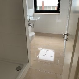 USŁUGI OGÓLNOBUDOWLANE RT - Nowoczesna, jasna łazienka z beżowymi płytkami podłogowymi, białą armaturą, kabiną prysznicową i oknem z czarną ramą.