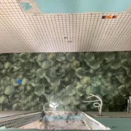 Łazienka w trakcie remontu: ściany i podłoga wyłożone heksagonalnymi płytkami w odcieniach zieleni, sufit z białych kwadratowych płytek z czarną fugą, widoczne przewody elektryczne i elementy...