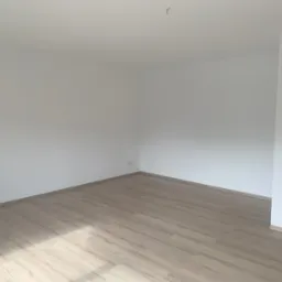 Pusty, odnowiony pokój z jasnymi panelami podłogowymi imitującymi drewno i świeżo pomalowanymi białymi ścianami, widoczne gniazdko elektryczne i wystający kabel ze ściany sufitu.