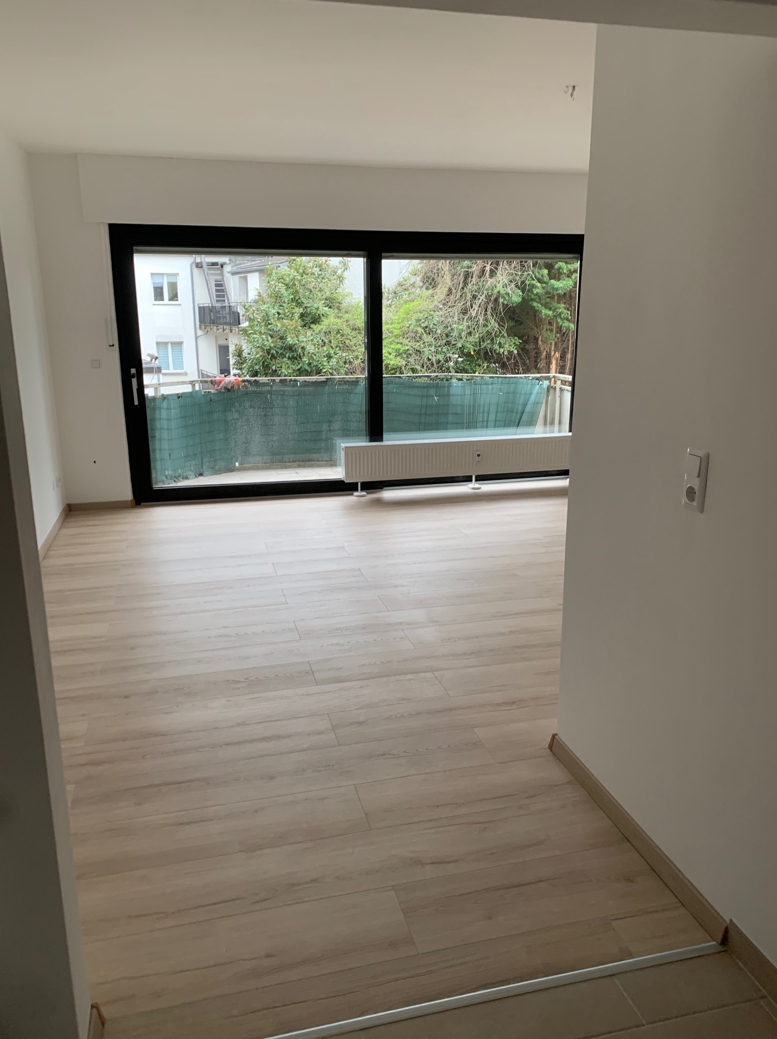 Pusty pokój z drewnopodobną podłogą, dużym oknem balkonowym z czarną ramą i grzejnikiem pod nim, widok z korytarza.