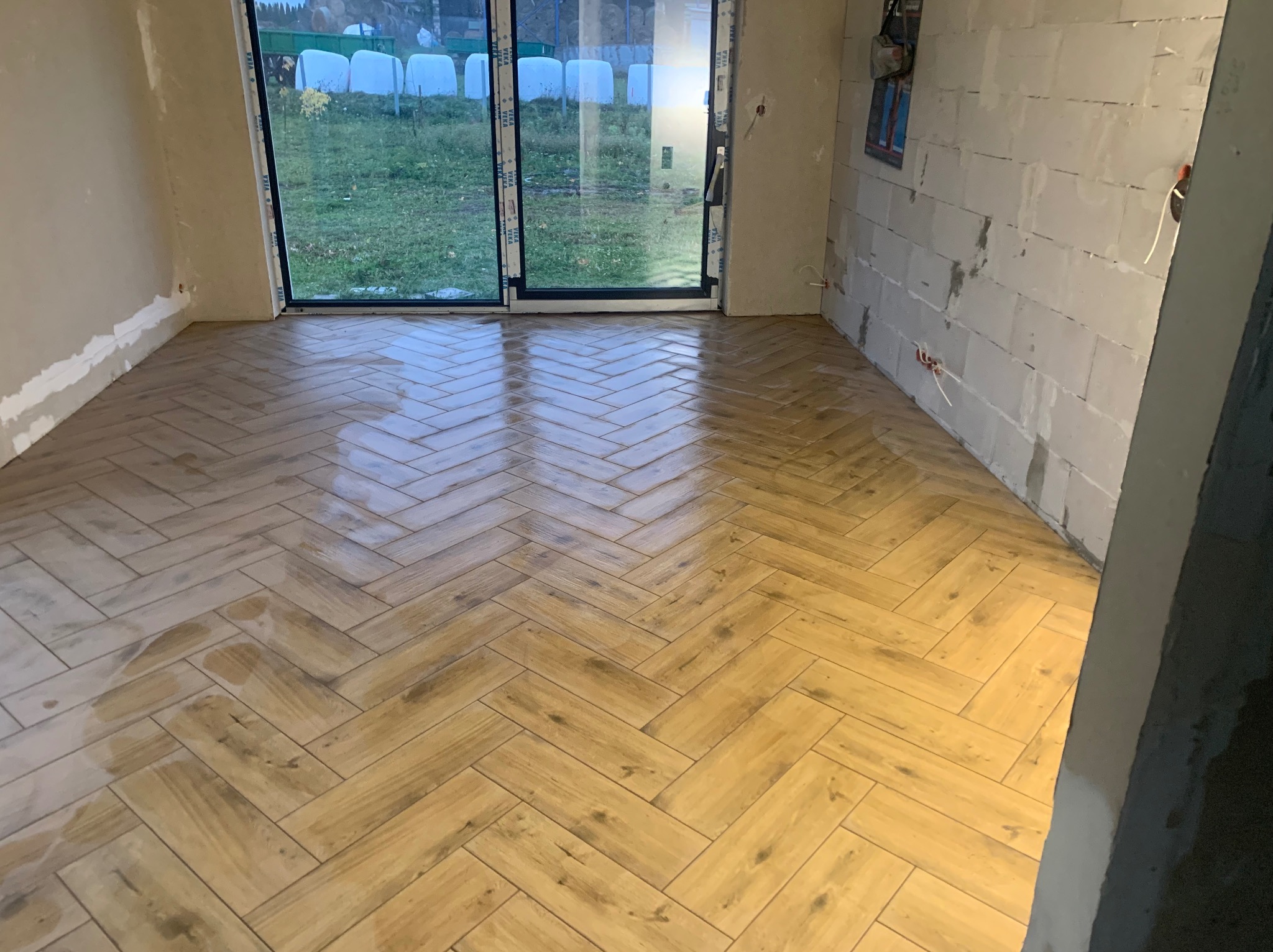 Ułożona pod kątem jasnobrązowa podłoga z płytek imitujących drewno w surowym pomieszczeniu z niezakończonymi ścianami i dużym oknem balkonowym.