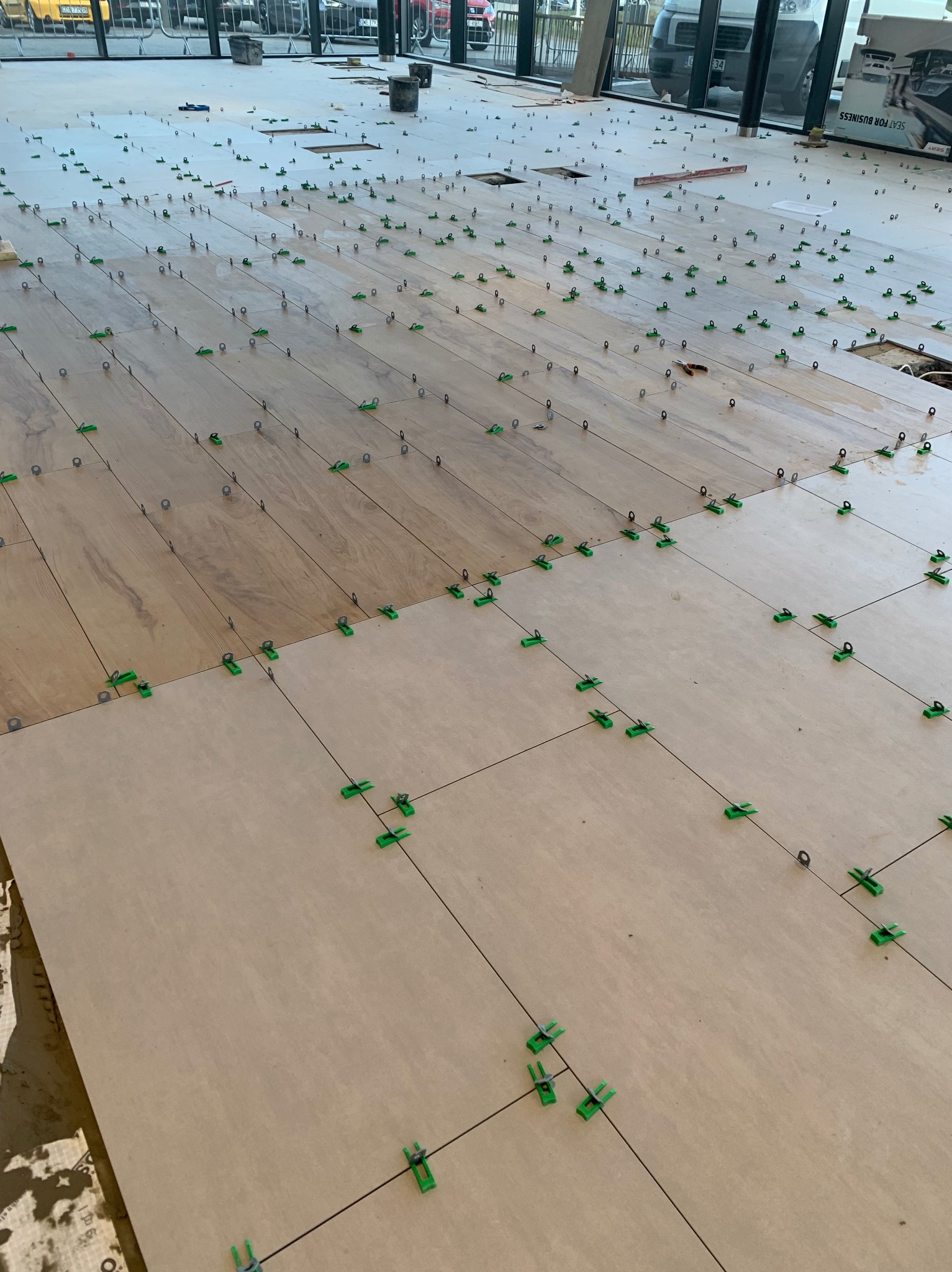 Układanie podłogi z płytek w dwóch kolorach, z użyciem systemu poziomowania z zielonymi klipsami i szarymi klinami, widoczne linie fug.