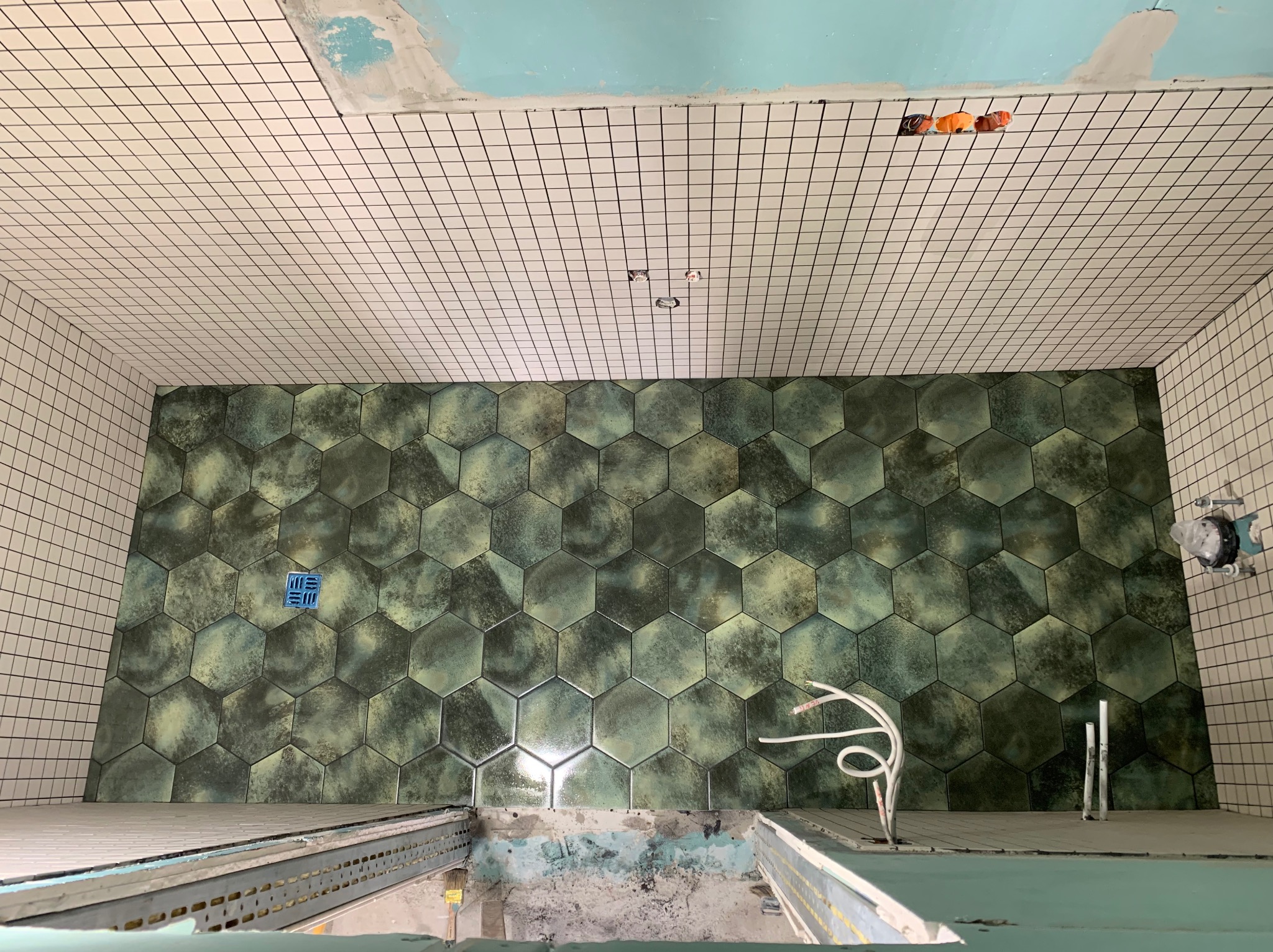 Łazienka w trakcie remontu: ściany i podłoga wyłożone heksagonalnymi płytkami w odcieniach zieleni, sufit z białych kwadratowych płytek z czarną fugą, widoczne przewody elektryczne i elementy...