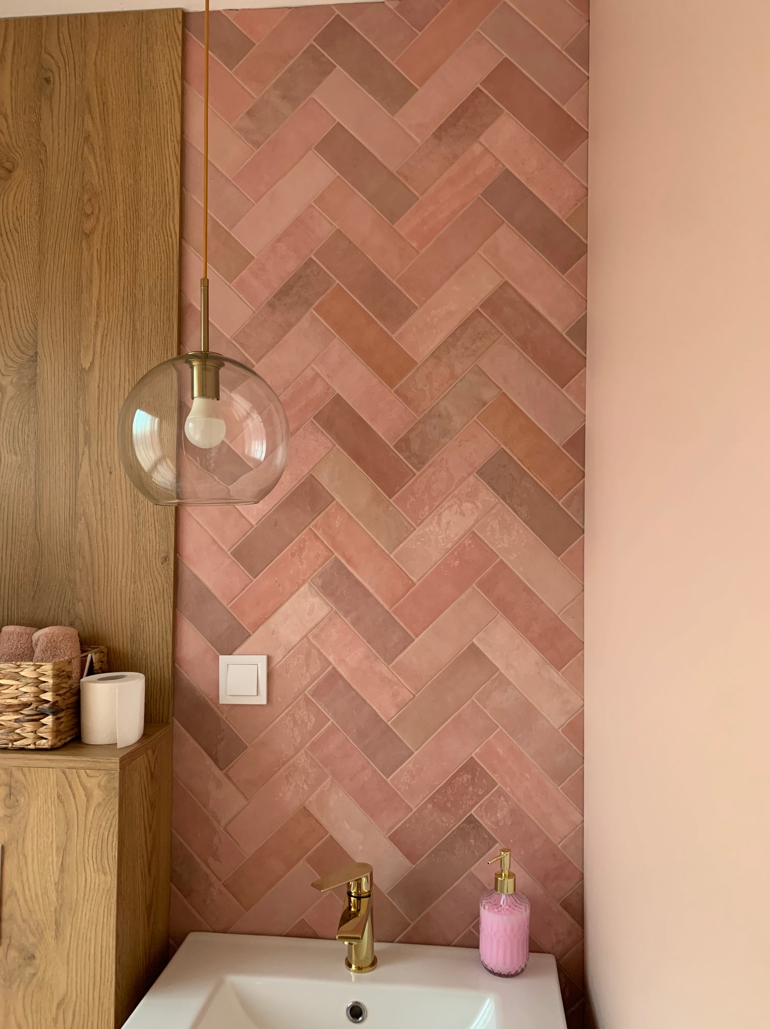 Łazienka z różową cegłą ułożoną w jodełkę, złota armatura, szafka z drewnianym fornirem i szklana lampa wisząca.
