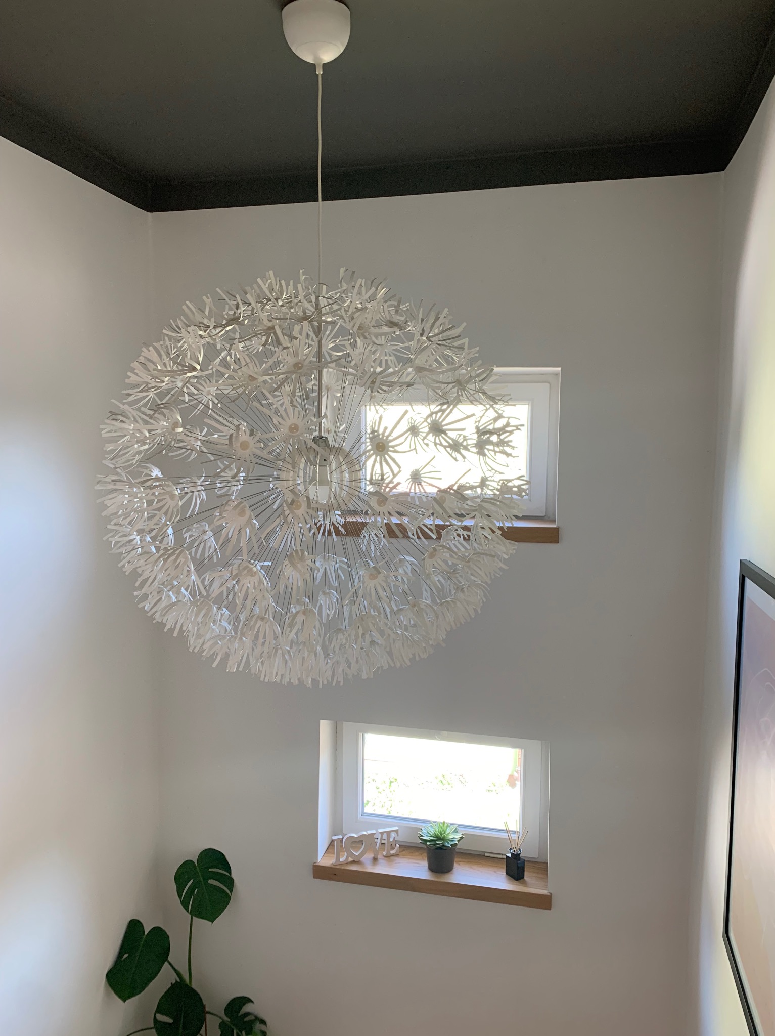 Wnętrze klatki schodowej z białą lampą w kształcie dmuchawca, czarnym sufitem, dwoma małymi oknami z drewnianymi parapetami i zieloną rośliną w doniczce.