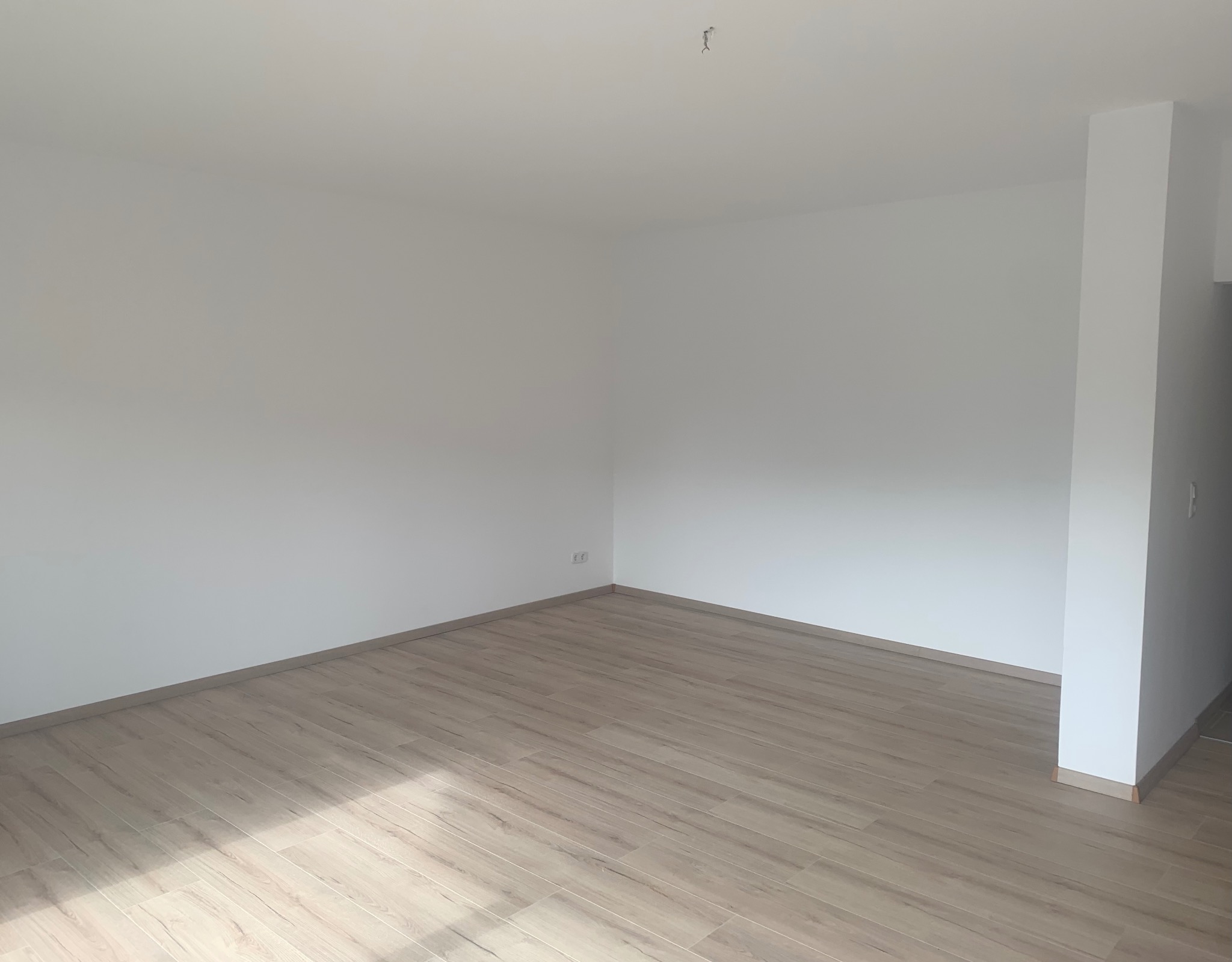 Pusty, odnowiony pokój z jasnymi panelami podłogowymi imitującymi drewno i świeżo pomalowanymi białymi ścianami, widoczne gniazdko elektryczne i wystający kabel ze ściany sufitu.
