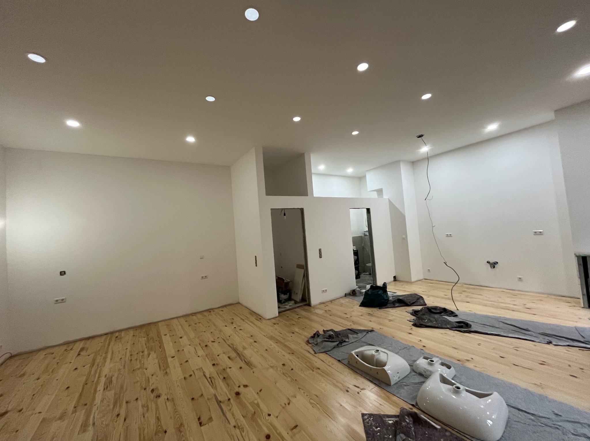 Wnętrze w trakcie prac wykończeniowych: świeżo pomalowane białe ściany, drewniana podłoga, przygotowane do montażu umywalki na podłodze zabezpieczonej folią, widoczne punkty oświetleniowe w suficie.