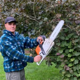 Mężczyzna w czapce i polarze w niebieską kratę trzyma piłę łańcuchową Stihl na tle krzewu z zielono-bordowymi liśćmi.