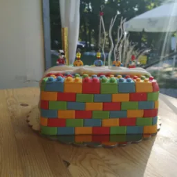 Tort urodzinowy w kształcie klocków, udekorowany figurkami i świeczkami, ustawiony na drewnianym stole.