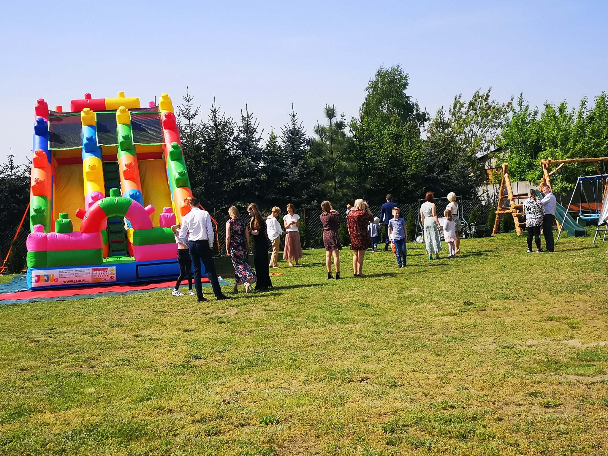 Kolorowa, dmuchana zjeżdżalnia na trawniku podczas imprezy plenerowej, z grupą osób w tle, w tym dzieci i dorośli, w słoneczny dzień.