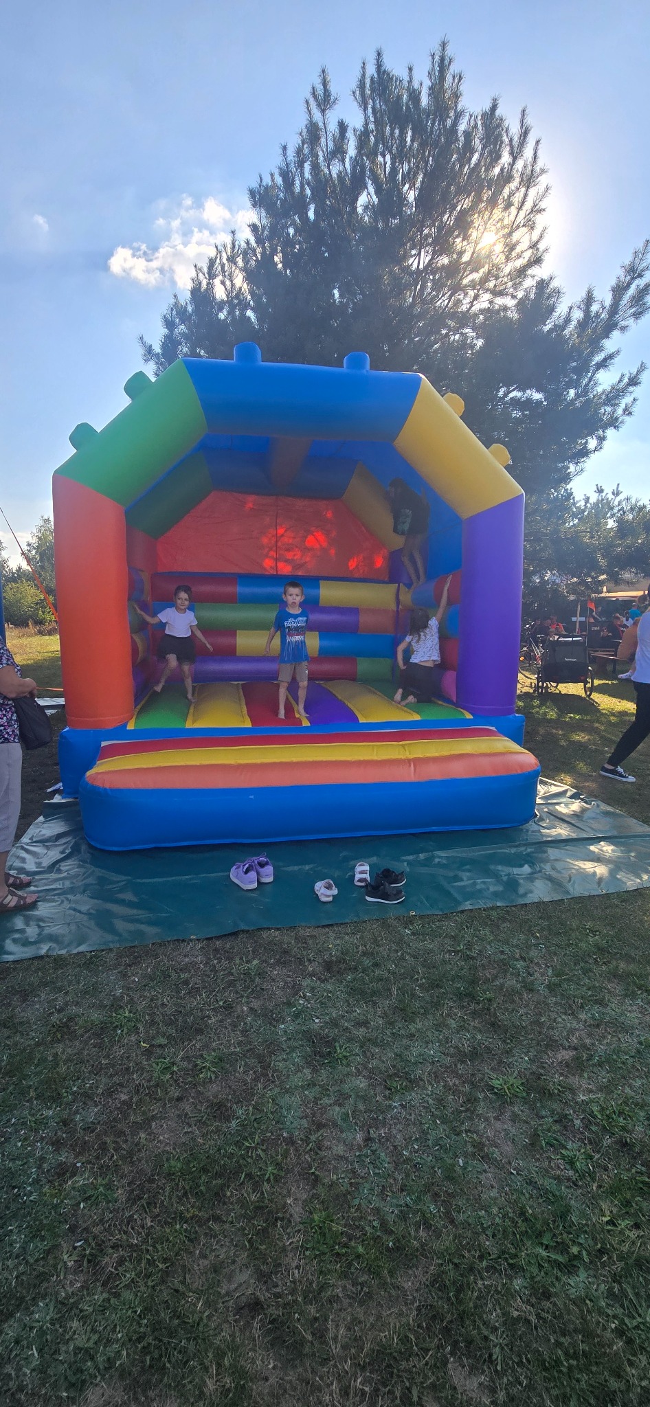 Kolorowy dmuchany zamek z dziećmi skaczącymi w środku, buty zdjęte i ułożone przed wejściem, na zielonej trawie w słoneczny dzień.