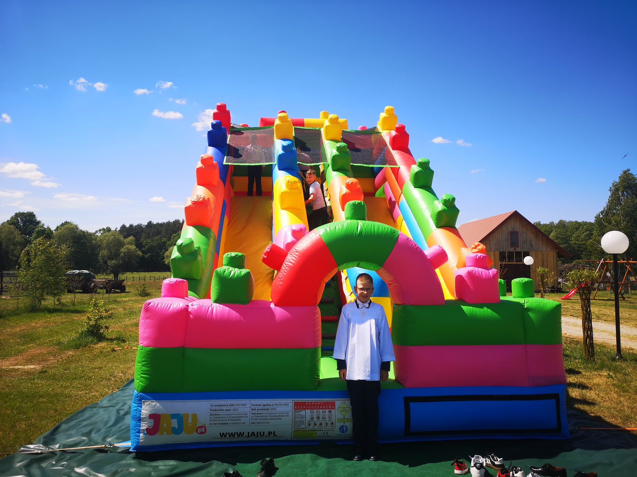 Chłopiec w stroju komunijnym stoi przed kolorowym, dmuchanym zamkiem ze zjeżdżalnią na trawiastym terenie z budynkiem w tle.