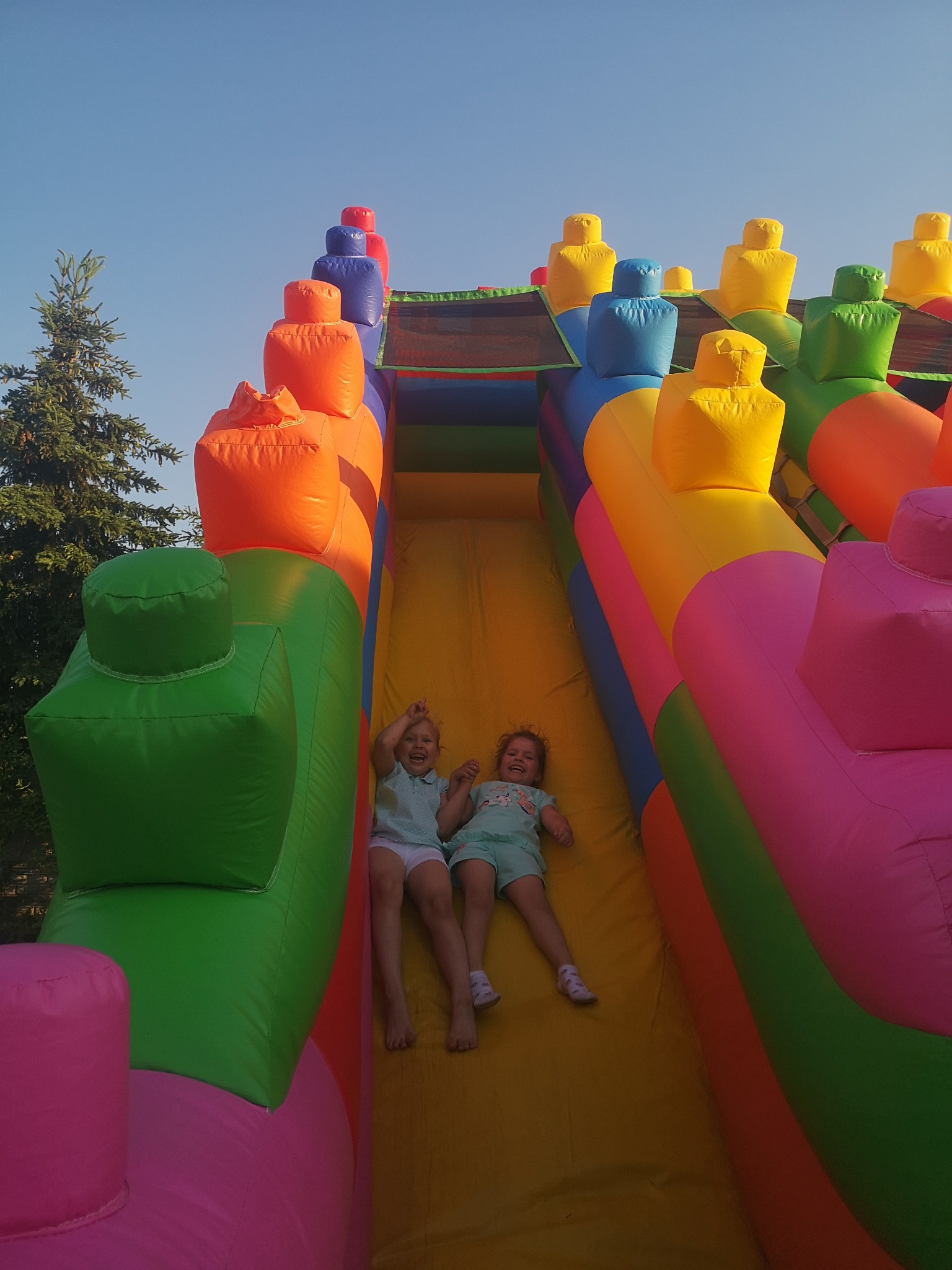 Dwie roześmiane dziewczynki zjeżdżają z kolorowej, dmuchanej zjeżdżalni w słoneczny dzień. Widoczna zielona choinka w tle.