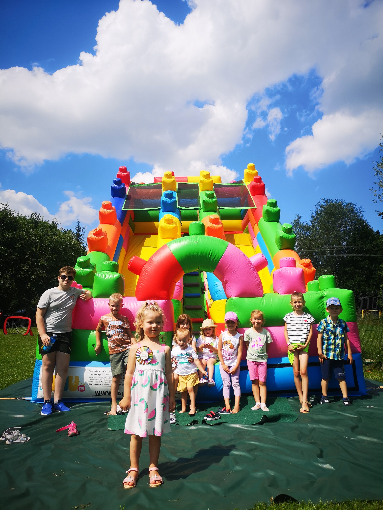 Grupa dzieci pozuje na tle kolorowego, dmuchanego zamku z przeszkodami na zielonej macie, w słoneczny dzień z błękitnym niebem i białymi chmurami.