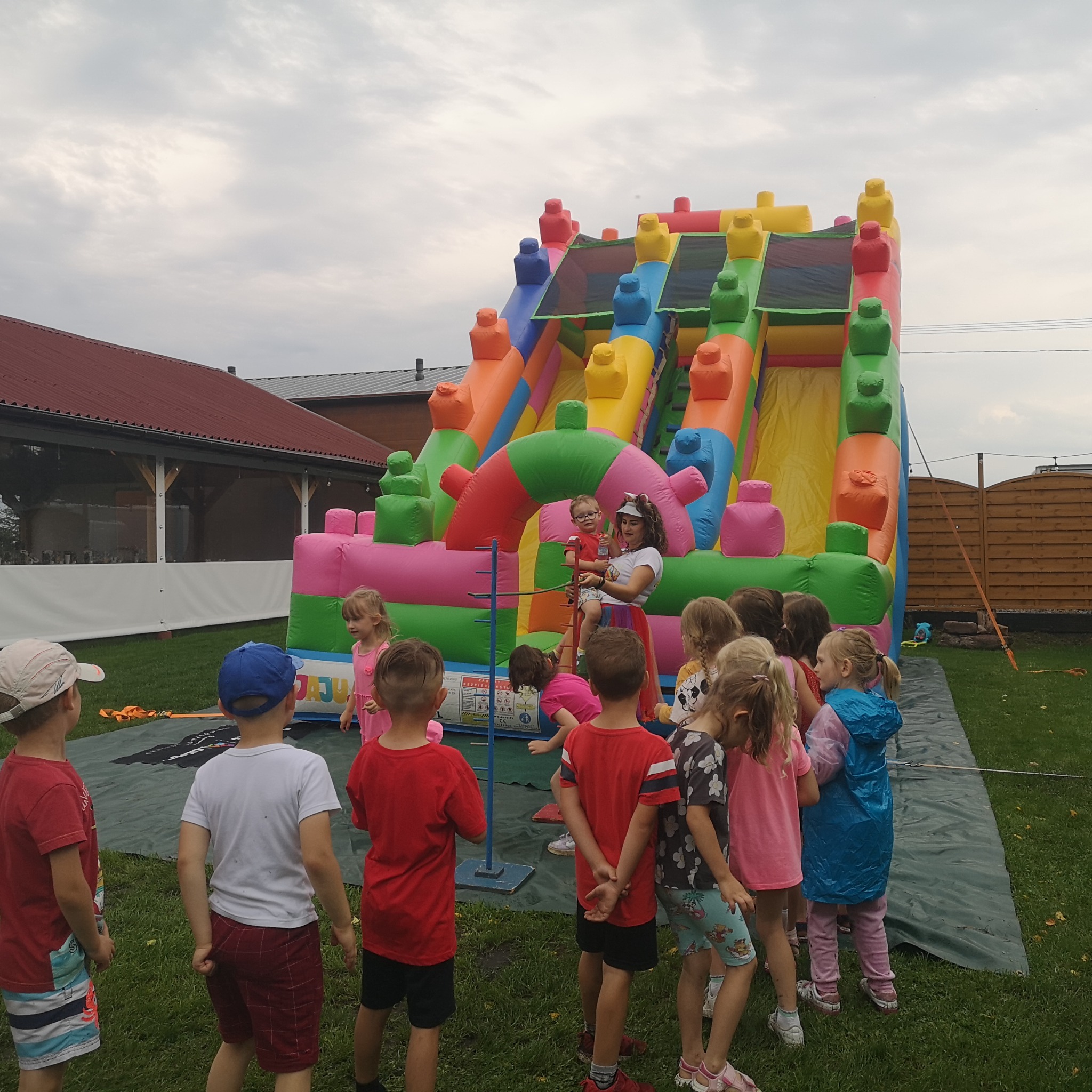 Dzieci zgromadzone przed dmuchaną zjeżdżalnią, animator trzyma na rękach małe dziecko, w tle budynek z czerwonym dachem i drewniany płot.