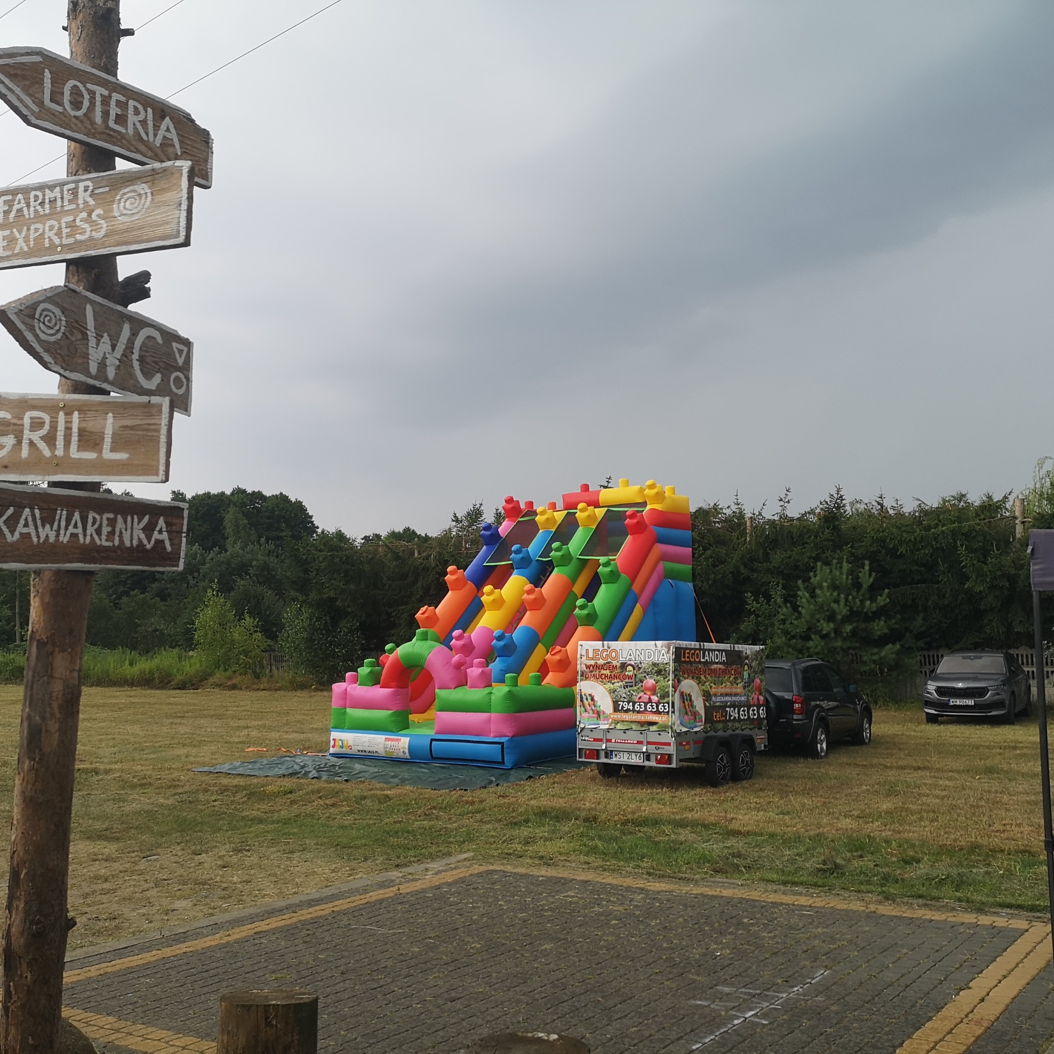 Kolorowy dmuchany zamek z przeszkodami, przypominający klocki Lego, ustawiony na trawie obok przyczepy z reklamą Legolandia, w tle drzewa i pochmurne niebo, w kadrze widoczny drewniany drogowskaz...