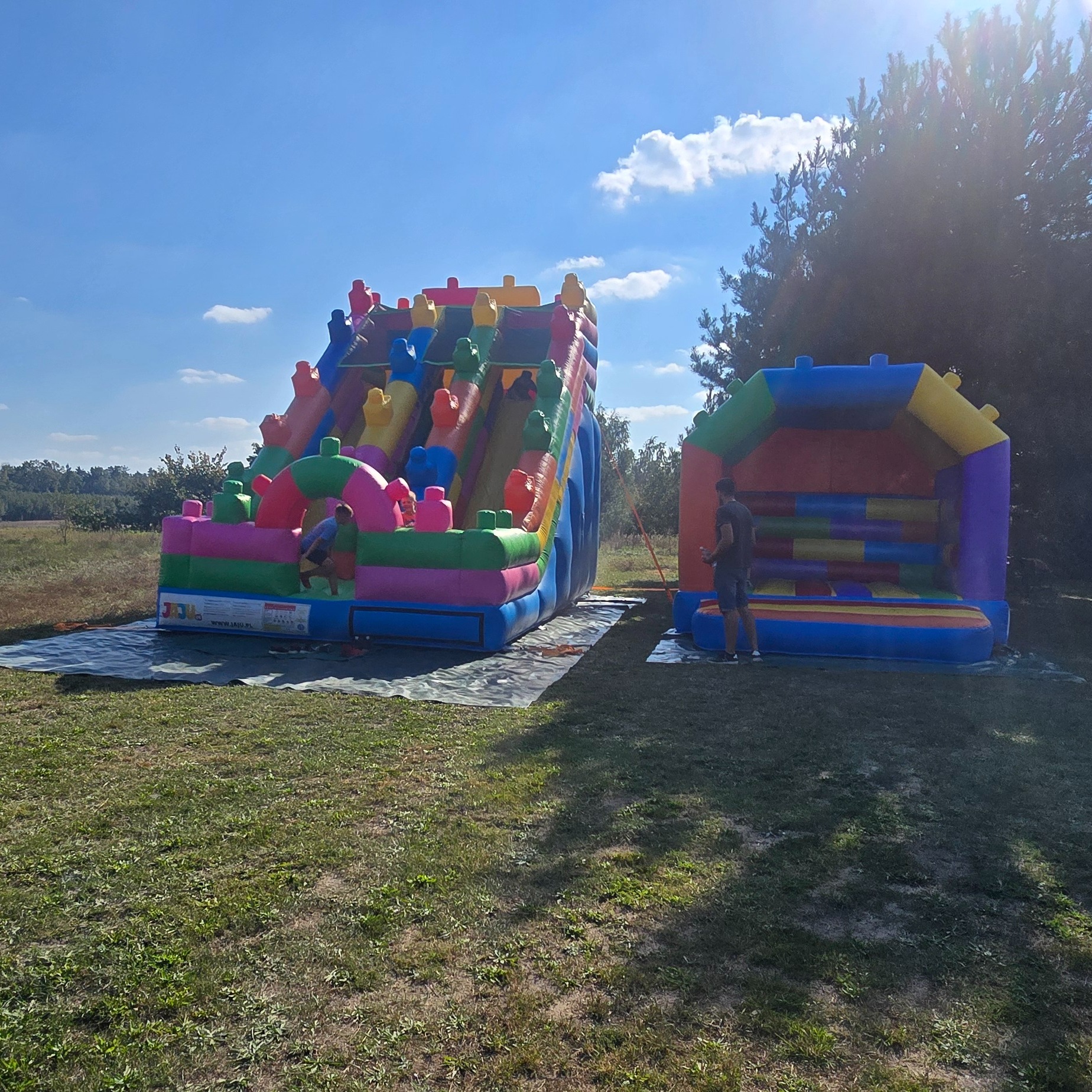 Kolorowa dmuchana zjeżdżalnia i zamek na trawie, częściowo rozłożone na folii ochronnej, z dziećmi w pobliżu, w słoneczny dzień.