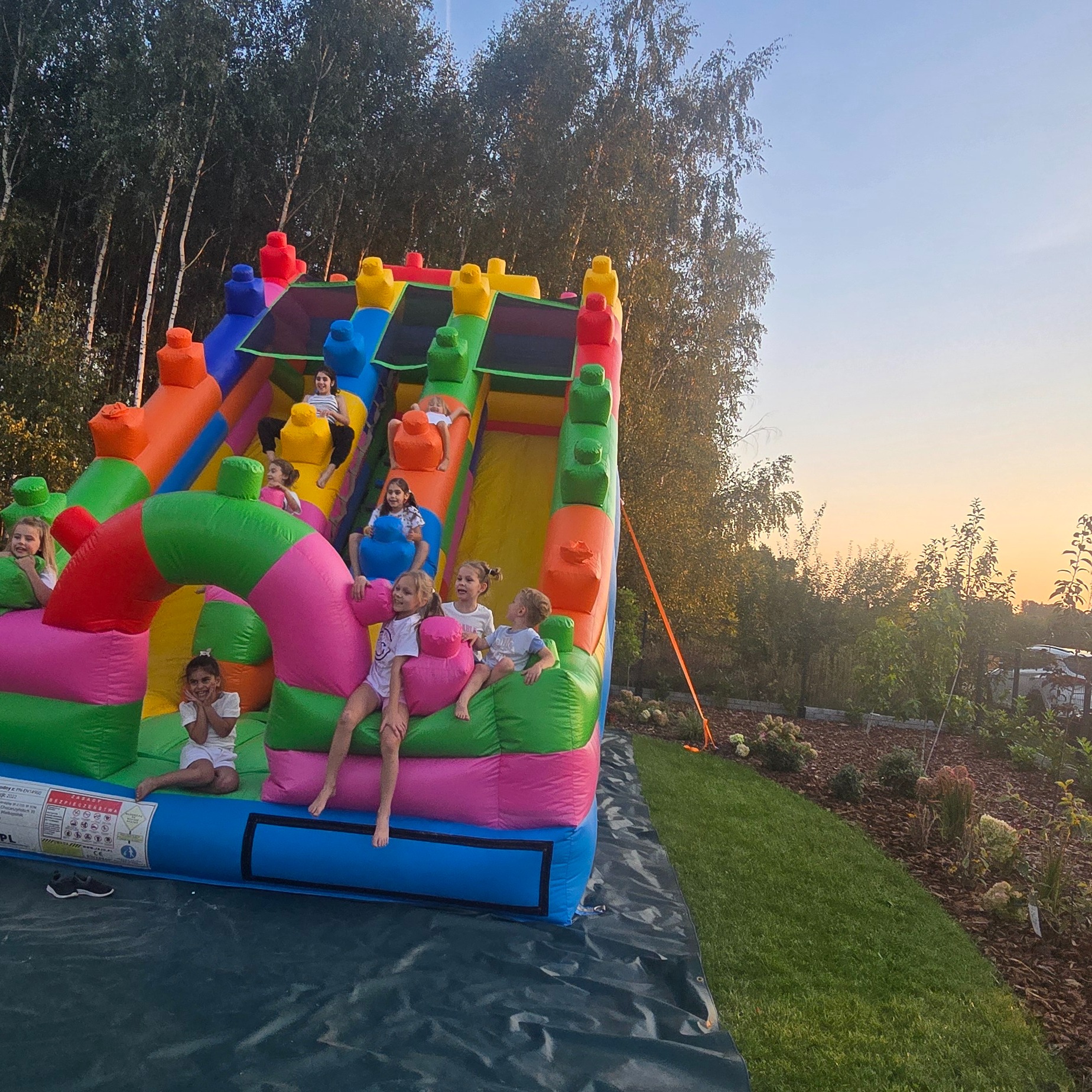 Kolorowa, dmuchana zjeżdżalnia z dziećmi bawiącymi się na niej, ustawiona na trawniku w ogrodzie o zmierzchu.