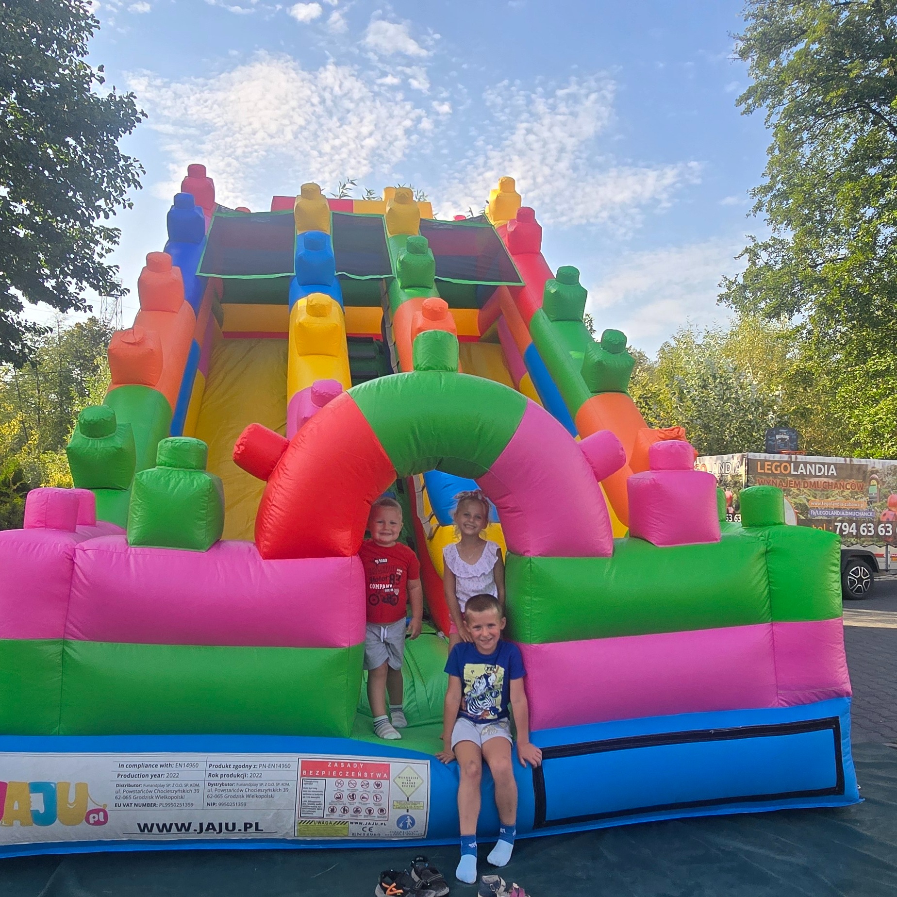 Trójka uśmiechniętych dzieci pozuje na tle kolorowej, dmuchanej zjeżdżalni z elementami w kształcie klocków, widoczny fragment reklamy 'Legolandia wynajem dmuchańców'.