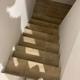 Wykończone schody z płytek imitujących drewno, widok z g&oacute;ry na stopnie i stopami w butach roboczych na dole schod&oacute;w.