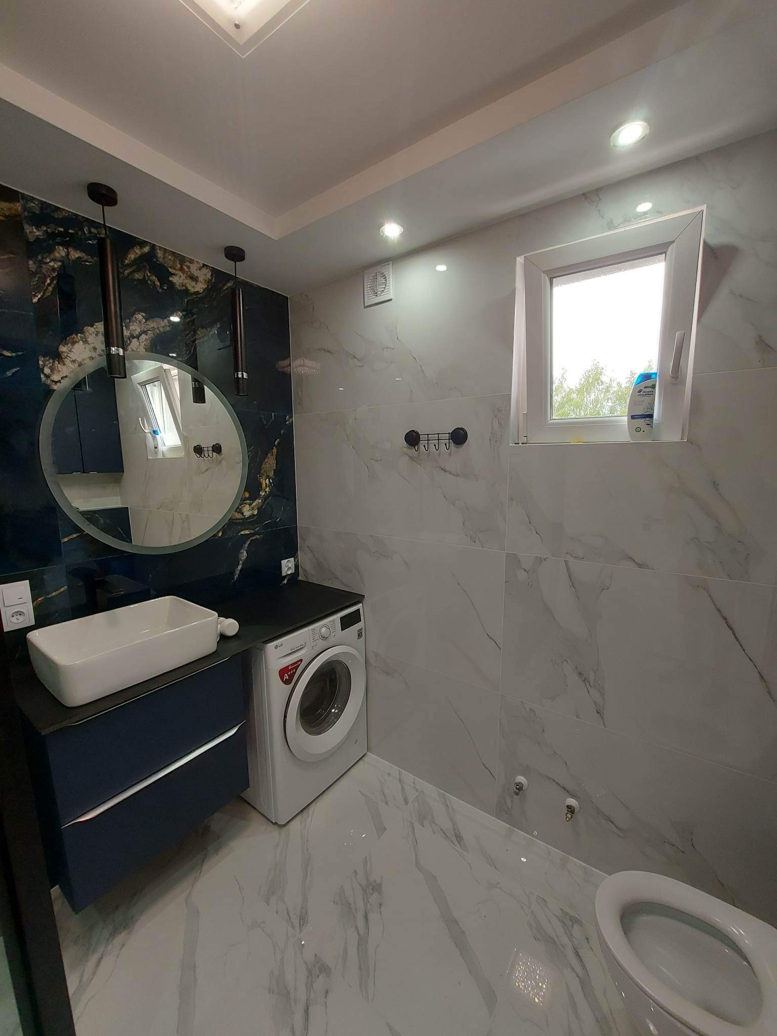 Nowoczesna łazienka z marmurowymi płytkami, umywalką nablatową na szafce w odcieniach granatu, okrągłym lustrem podwieszonym na tle ściany z efektem marmuru i pralką pod blatem, oświetlona...