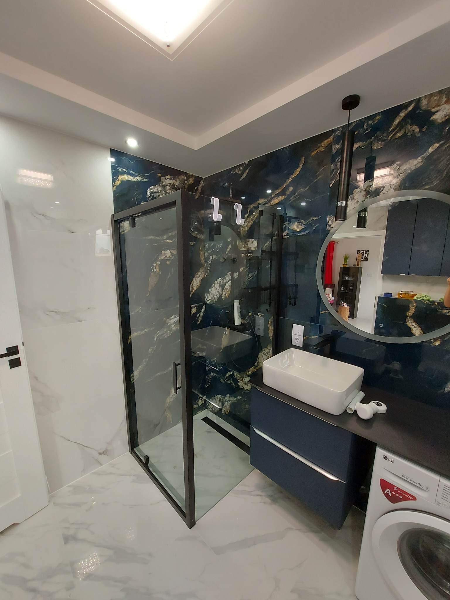 Nowoczesna łazienka z kabiną prysznicową typu walk-in, umywalką nablatową na szafce w kolorze granatowym, marmurowymi płytkami w odcieniach bieli i granatu oraz pralką LG z przodu.