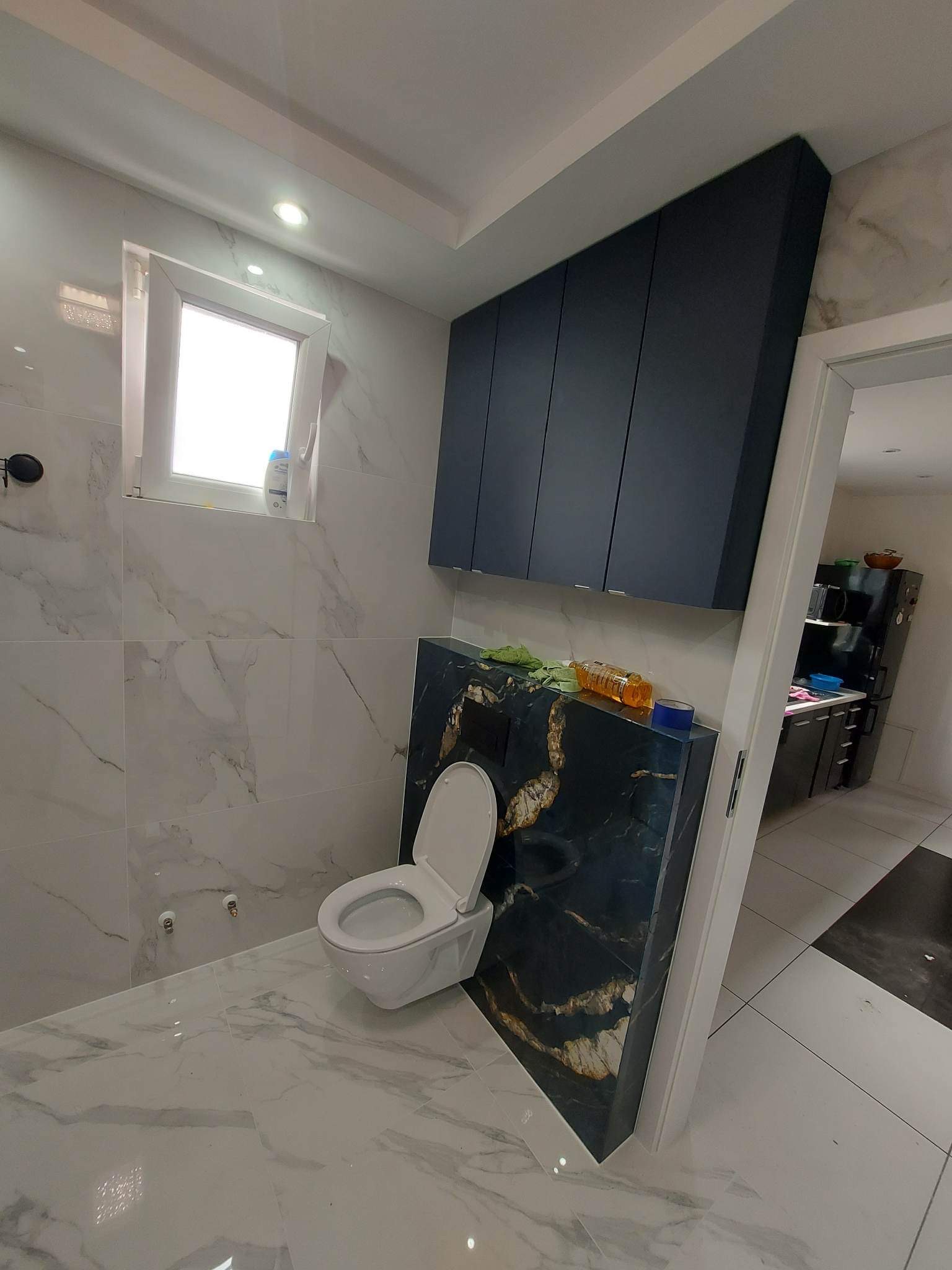 Nowoczesna łazienka z marmurowymi płytkami, wiszącą toaletą z czarnym panelem dekoracyjnym z akcentami złota, oraz ciemnoniebieskimi szafkami wiszącymi.