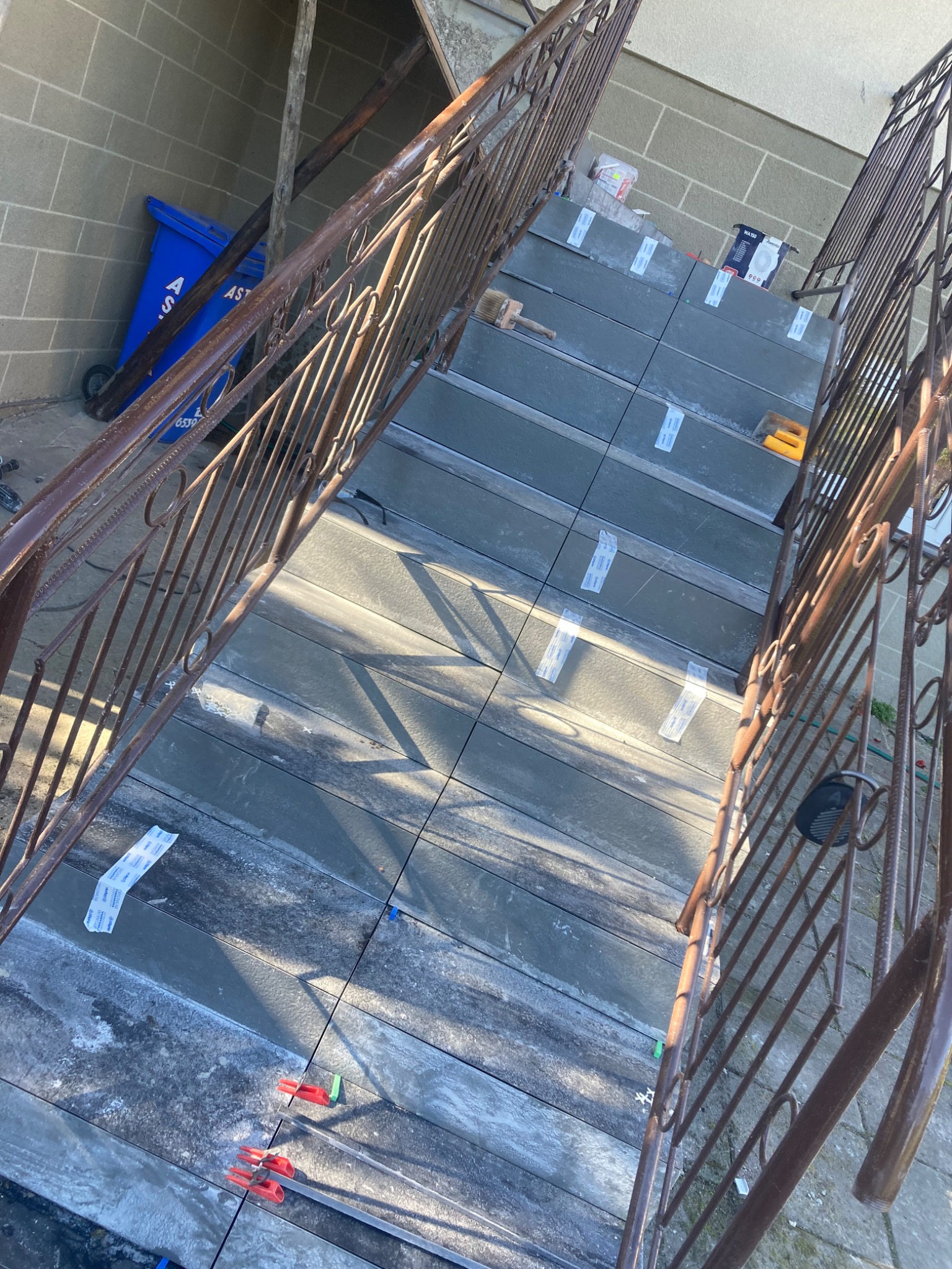 Kładzenie płytek na schodach zewnętrznych, widoczne listwy dystansowe i poziomica, metalowa balustrada po bokach, w tle śmietnik i fragment ściany.