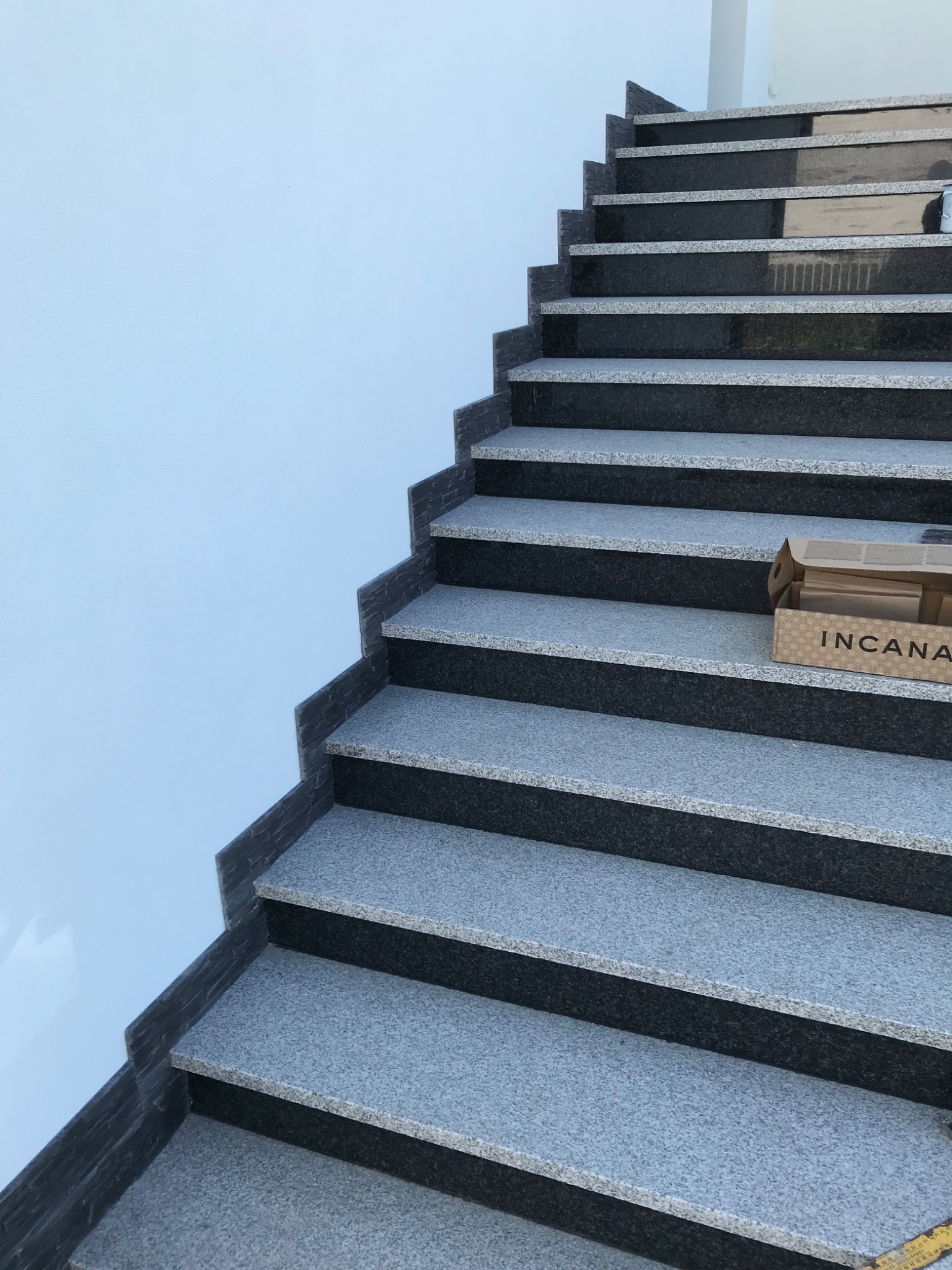 Schody z szarego granitu z czarnymi podstopnicami, przy białej ścianie, z pudełkiem płytek dekoracyjnych Incana na jednym ze stopni.