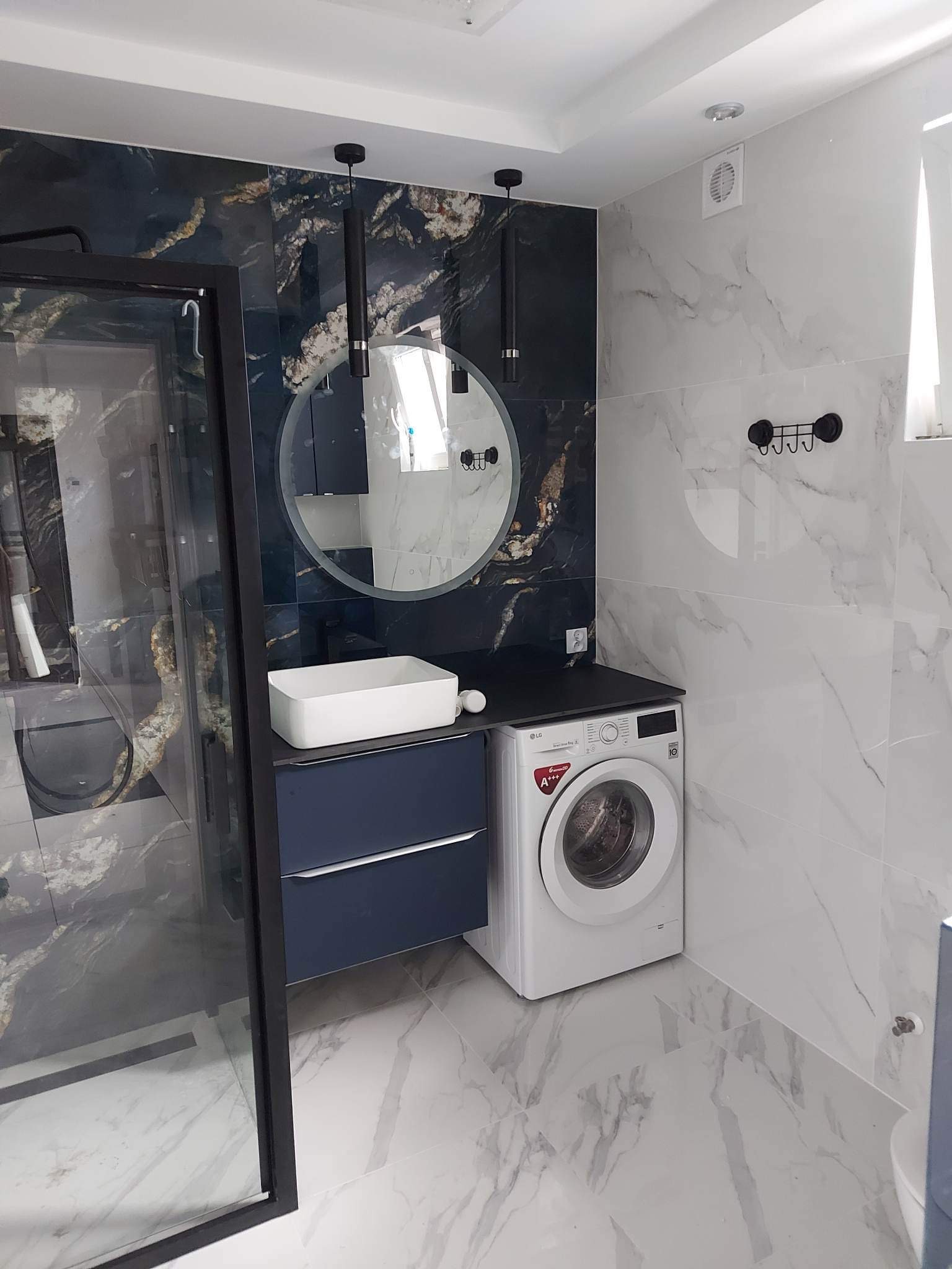 Nowoczesna łazienka z marmurowymi płytkami, kabiną prysznicową z czarną ramą, okrągłym lustrem z podświetleniem, umywalką nablatową na szafce w kolorze granatowym i pralką.