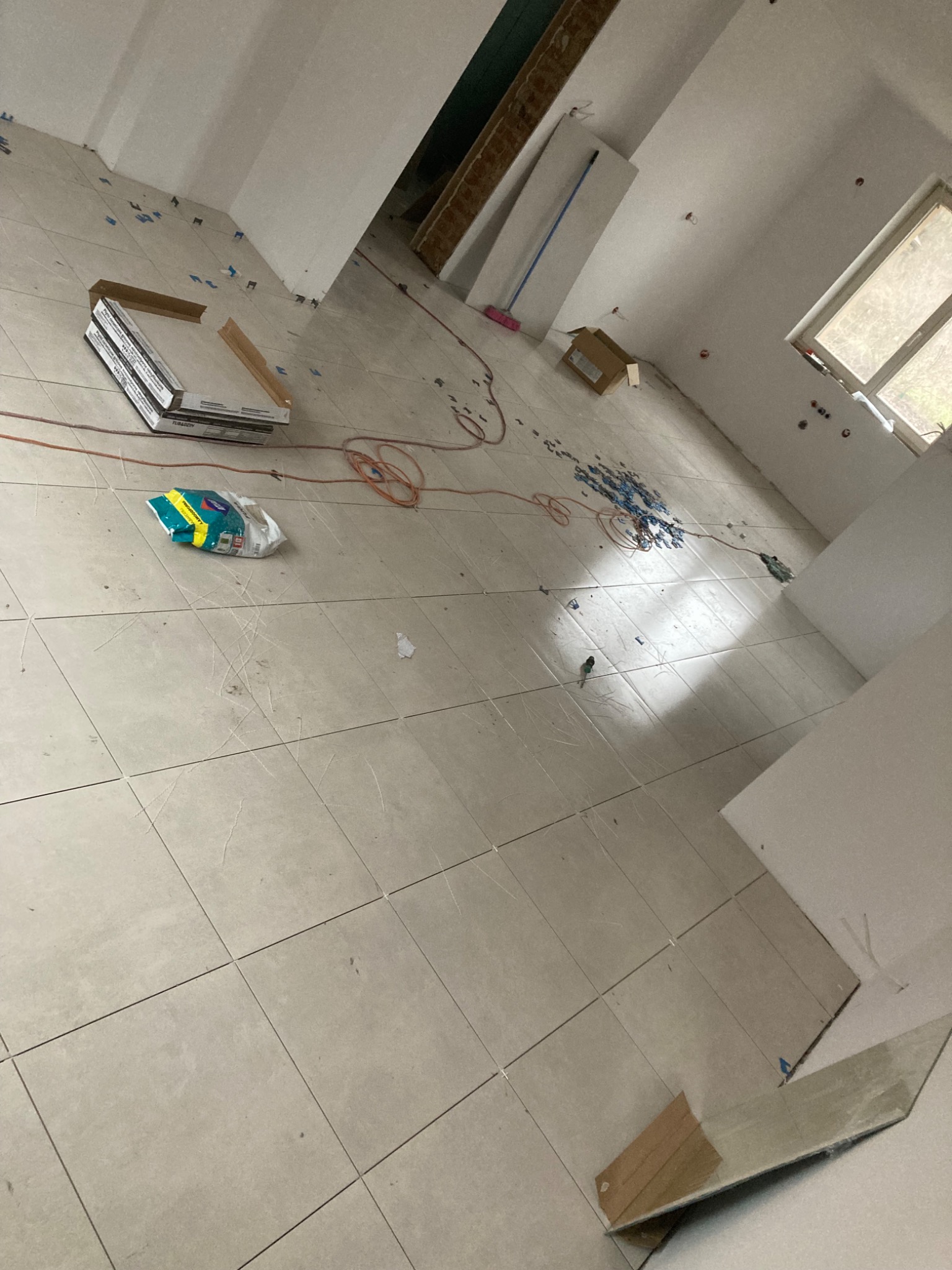 Widok na wnętrze w trakcie remontu: ułożona podłoga z płytek, paczka płytek, poziomica, niebieskie krzyżyki dystansowe, przewód elektryczny, karton i ślady po zaprawie na płytkach.