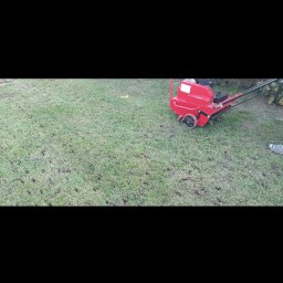 Konserwacja terenów zielonych - Aeracja trawnika: czerwony aerator na zielonej trawie, widoczne ślady po nakłuciach, fragment buta osoby obsługującej urządzenie w prawym dolnym rogu.