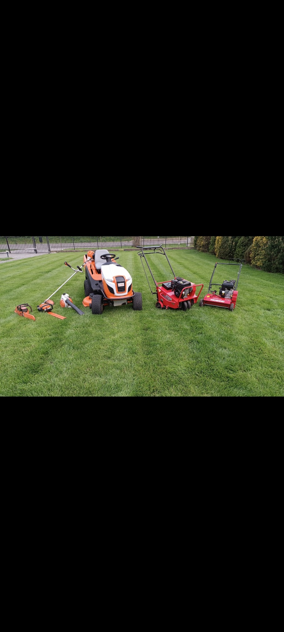 Różne maszyny ogrodowe: traktorek, wertykulator, kosiarka i dmuchawa, ułożone na świeżo skoszonej trawie z widocznymi pasami. Prezentacja sprzętu ogrodniczego.