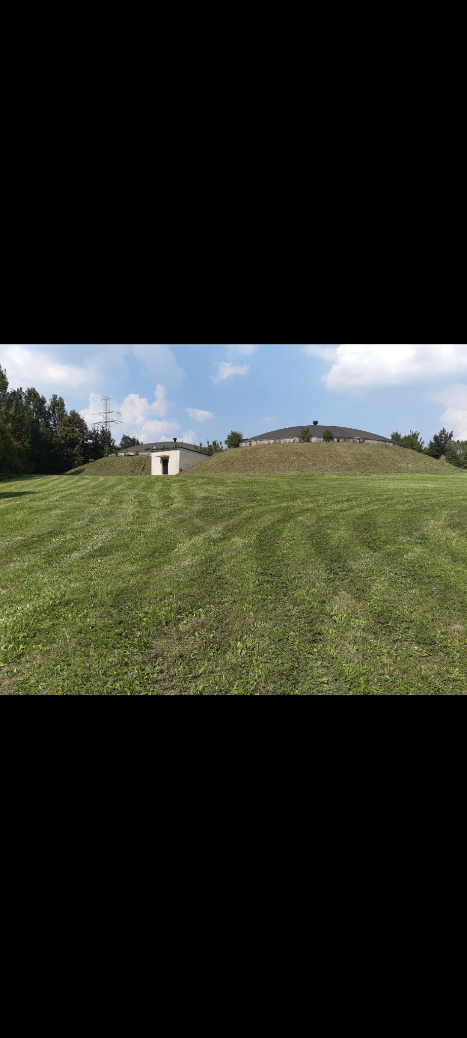 Rozległy trawnik z równo przystrzyżoną trawą, na wzgórzu widoczne budowle z płaskimi dachami. W tle drzewa i słupy wysokiego napięcia pod błękitnym niebem.