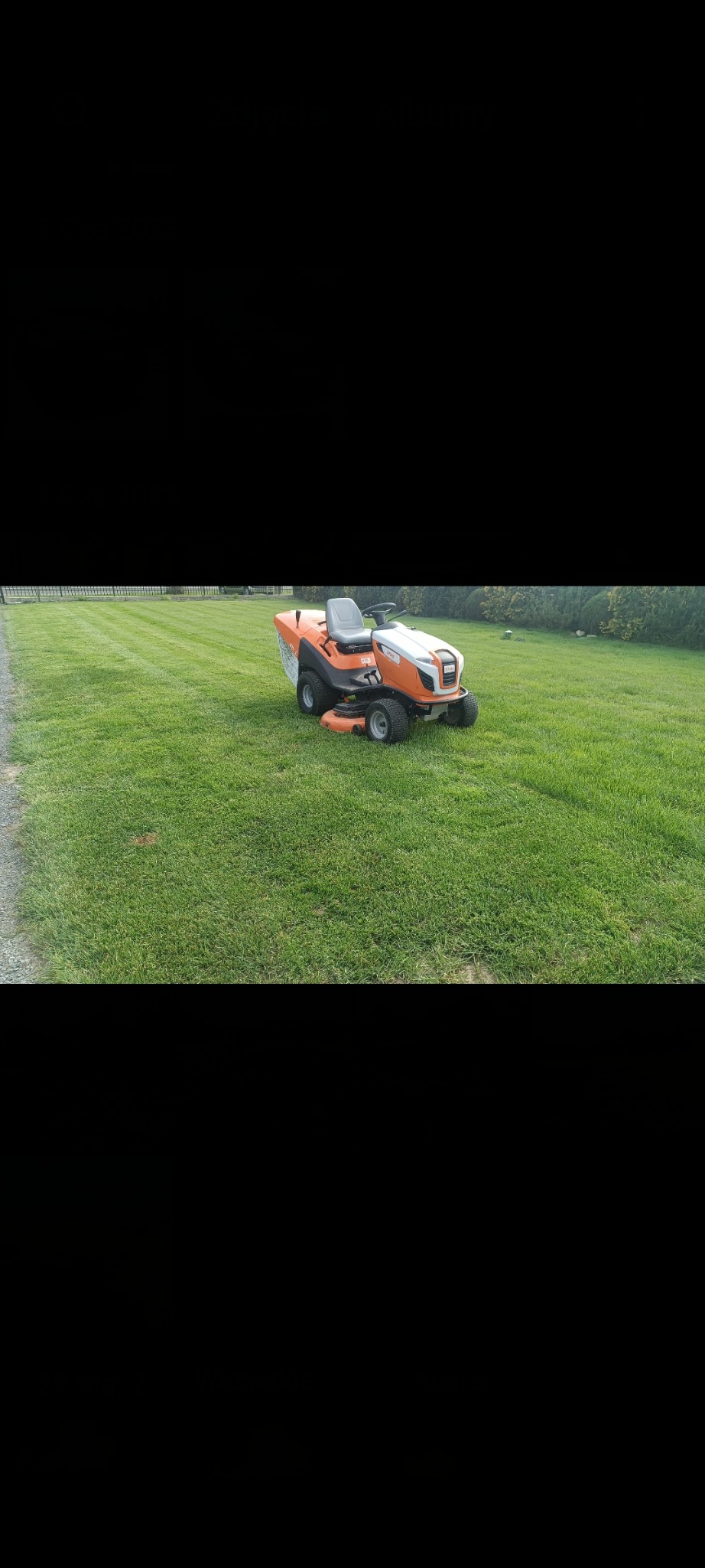 Pomarańczowa kosiarka traktorek na idealnie przystrzyżonym trawniku z widocznymi pasami, w tle żywopłot i drzewa. Słoneczny dzień.