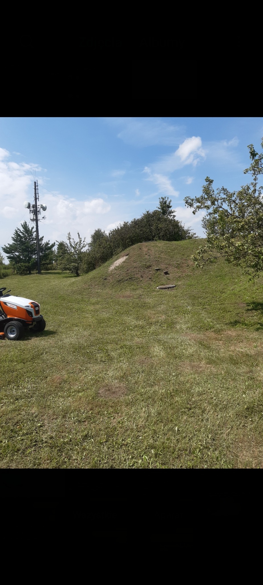 Pomarańczowy traktorek do trawy na nierównym terenie z pagórkiem i drzewami w tle, pod błękitnym niebem. Widoczna wieża telekomunikacyjna.