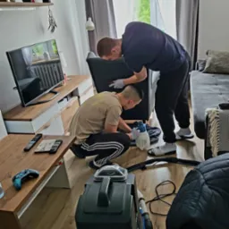 Dw&oacute;ch mężczyzn w rękawicach ochronnych czyści ciemnoszarą tapicerkę fotela za pomocą profesjonalnego odkurzacza piorącego i spryskiwacza w jasnym salonie.