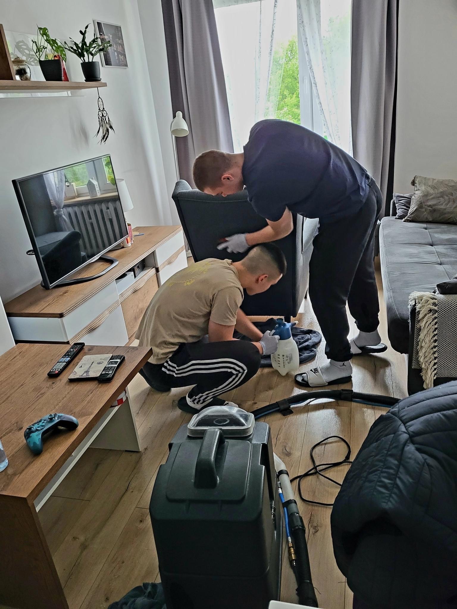 Dwóch mężczyzn w rękawicach ochronnych czyści ciemnoszarą tapicerkę fotela za pomocą profesjonalnego odkurzacza piorącego i spryskiwacza w jasnym salonie.