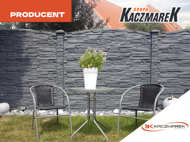 Szary ogrodzenie betonowe o fakturze łupka z dekoracyjnym wykończeniem, w tle dom z czerwoną dachówką, na pierwszym planie stolik kawowy z dwoma krzesłami i doniczką z kwiatami.