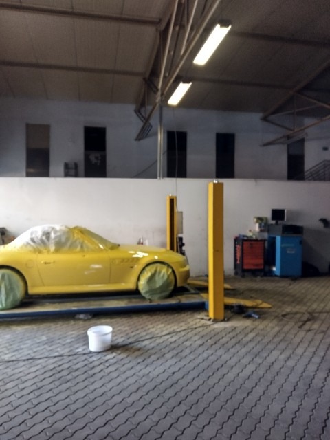 Żółty samochód sportowy na podnośniku warsztatowym, częściowo zakryty folią ochronną i zabezpieczonymi kołami, przygotowany do lakierowania. Białe wiadro na podłodze warsztatu.